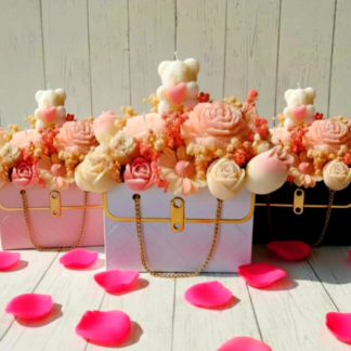 Trois bouquets de bougies gourmandes en cire d'olive pour la Saint-Valentin, présentés dans des sacs à main rose, blanc et noir, décorés de fleurs en cire et d'un ourson sur un fond boisé clair.